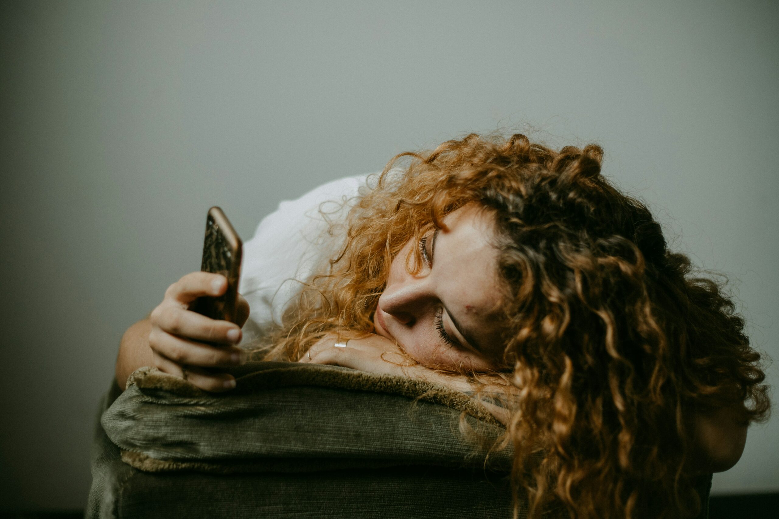 A woman with curly hair lies on a couch, staring at her phone with a distant, pained expression—capturing the loneliness, sadness, and emotional exhaustion often felt after a breakup.