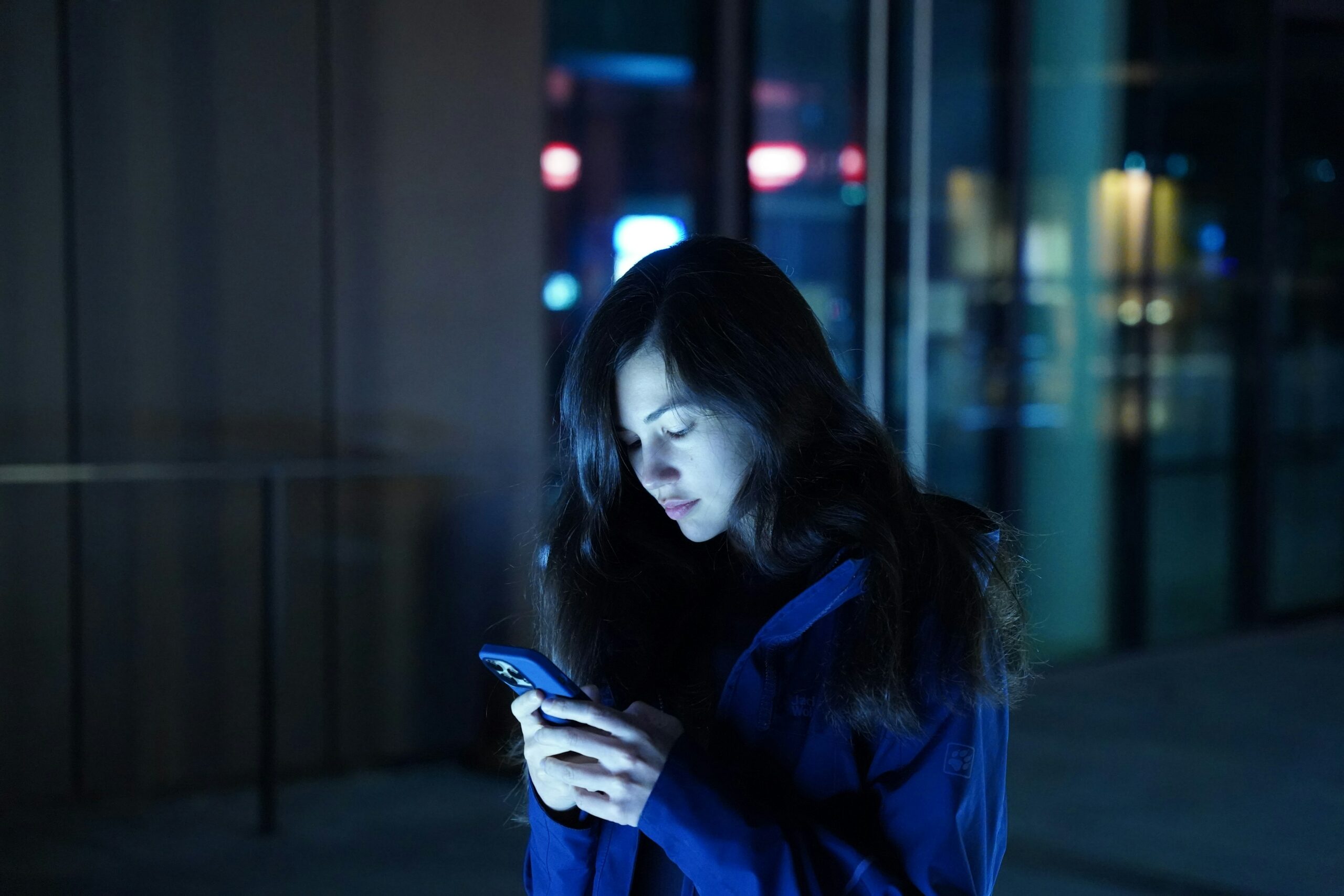 Woman standing alone at night, looking down at her phone, lit by blue light.